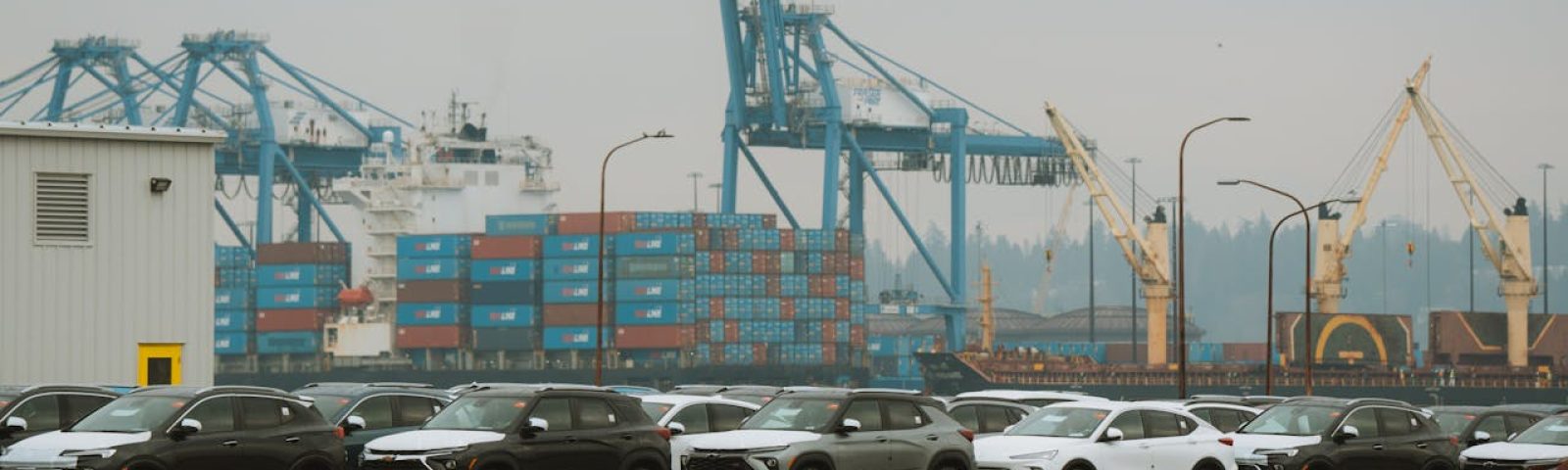 A lineup of parked cars at a bustling industrial shipping port with cranes and containers.